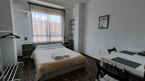 Habitación de hotel con cama, escritorio y silla. Se ve una ventana con cortinas, un perchero y una estantería con una lámpara.