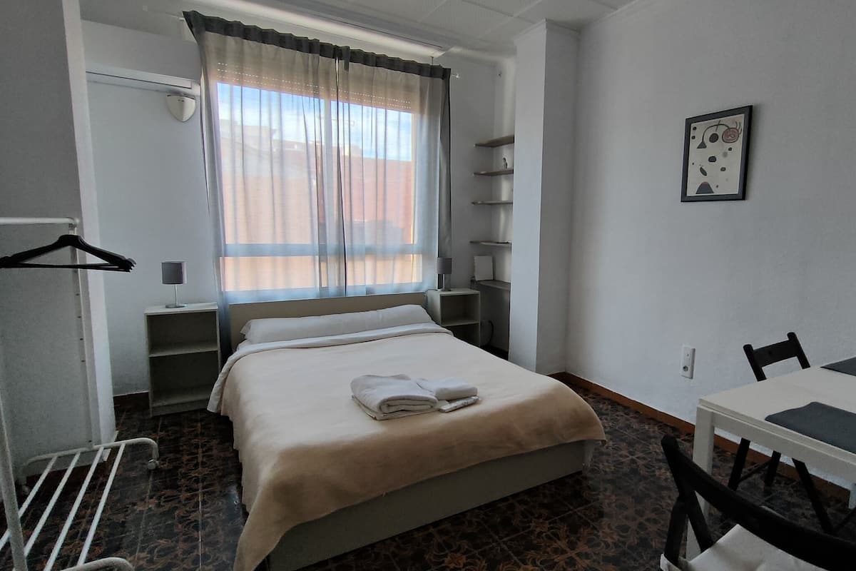 A hotel room with a bed, a desk, and a chair. There is a window with curtains, a coat rack, and a shelf with a lamp.