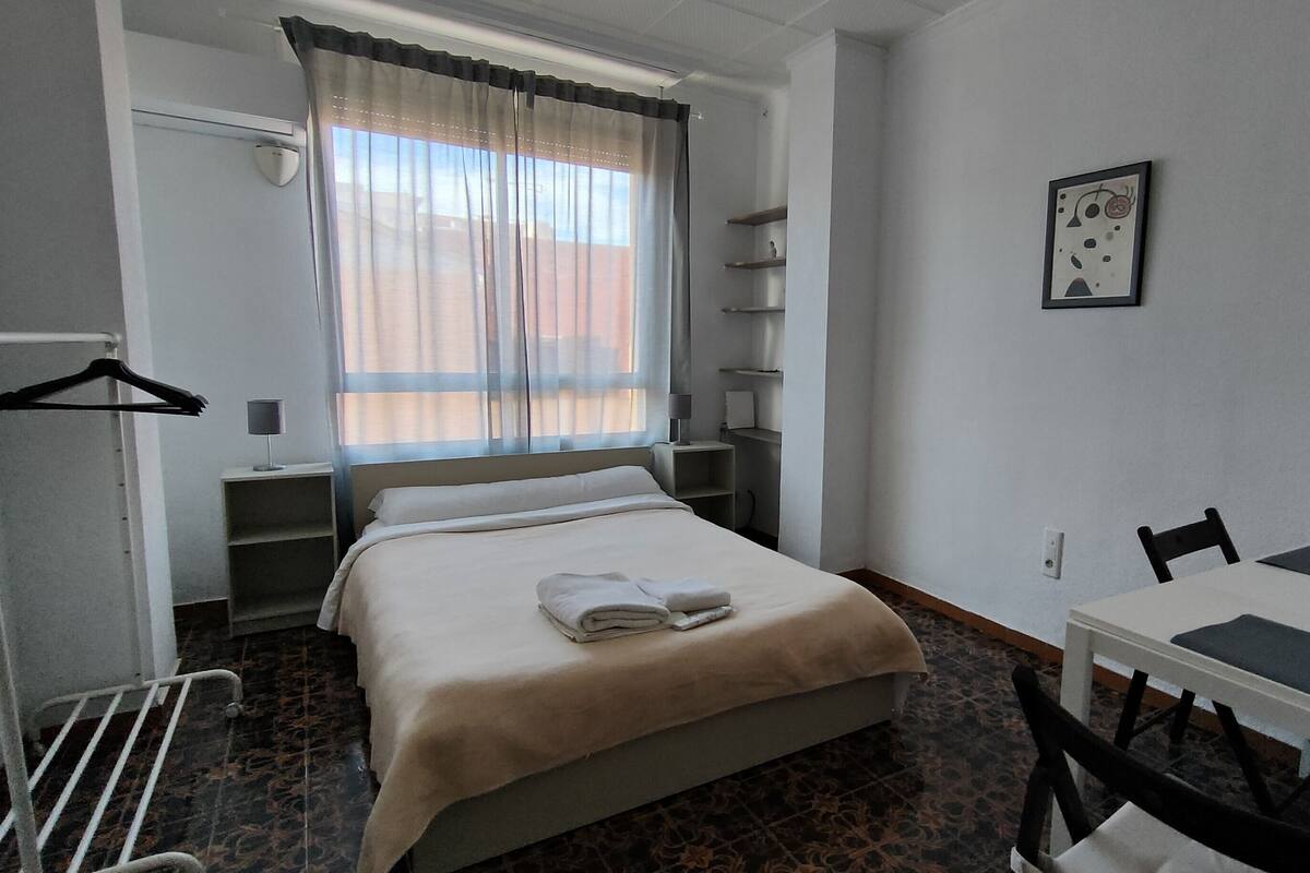 A hotel room with a bed, a table, chairs, a desk, a hanging coat rack, and a window with curtains.