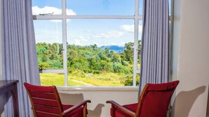 A room with a large window offering a view of a green landscape and mountains. There are two red chairs and a small table. - Brondesbury Park Hotel (Juliasdale)