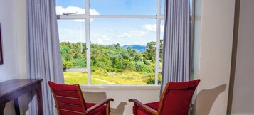Habitación con ventana grande que ofrece vistas a un paisaje verde y montañas. Hay dos sillas rojas y una mesita.