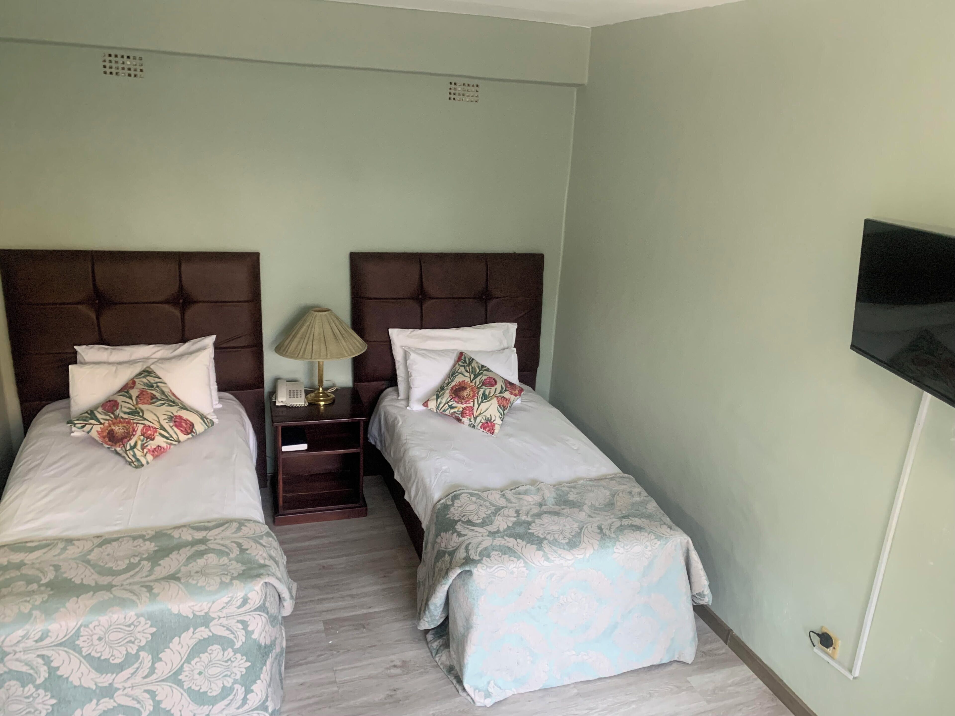 A hotel room with two single beds, a nightstand with a lamp, a TV mounted on the wall, and a light-colored floor.