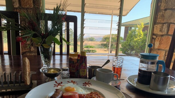A breakfast plate with sausage, tomato, beans, and fried plantains, a coffee pot, condiments, and a glass of iced tea on a table with a view of the outdoors. - Brondesbury Park Hotel (Juliasdale)