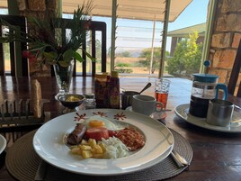 Un plato de desayuno con salchicha, tomate, frijoles y tajadas de plátano frito, una cafetera, aderezos y un vaso de té helado sobre una mesa con vistas al exterior.