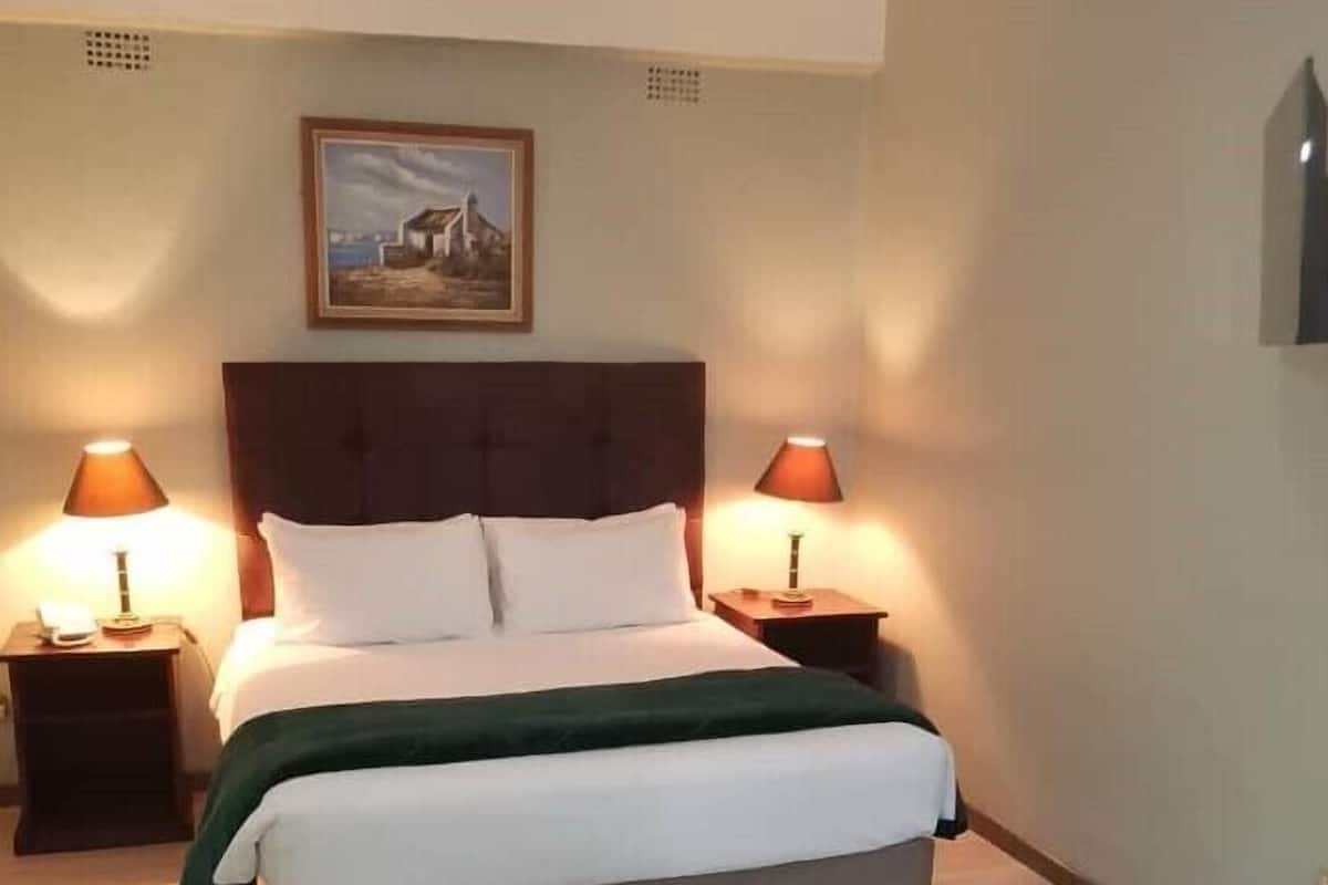 Un lit bien fait, agrémenté d’un plaid vert, deux lampes de chevet et un tableau encadré au mur.