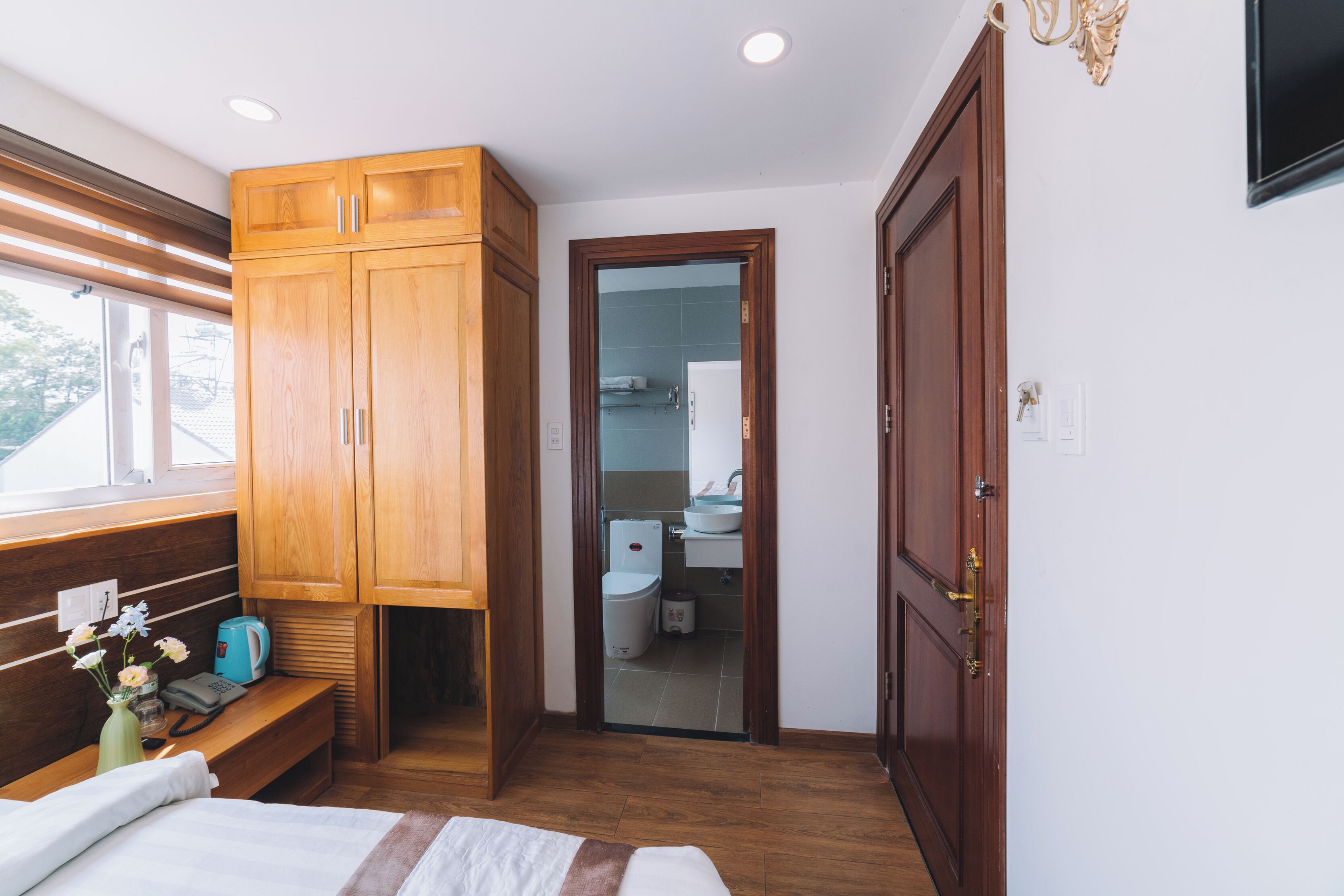 A bedroom with wooden cabinets, a bed, a bedside table with a vase and a telephone, and a view of the bathroom through an open door.