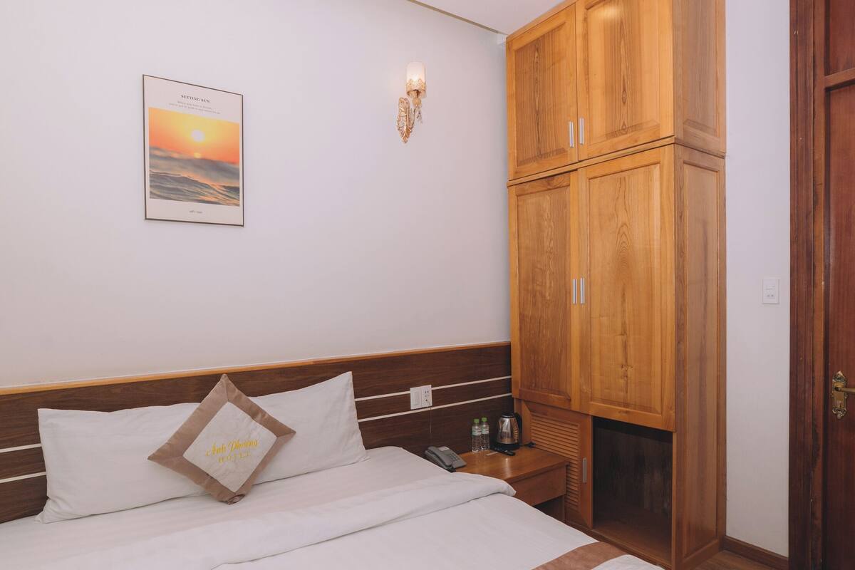 Een net opgemaakt bed met hoofdbord, een houten kledingkast, een ingelijst beeld aan de muur en een deur aan de rechterkant.