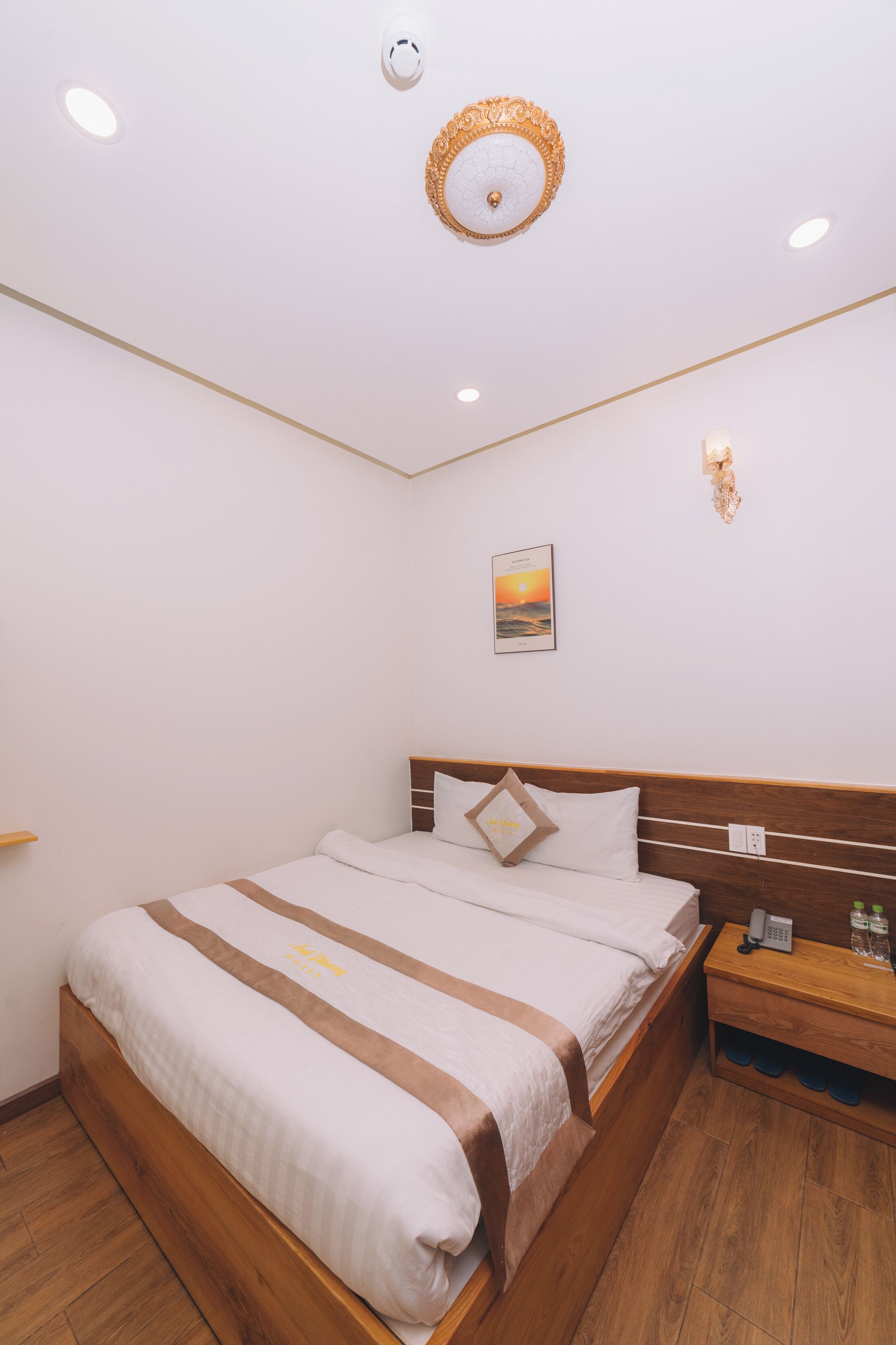 A neatly arranged hotel room with a wooden bed, a bedside table with a phone and toiletries, and a wall-mounted light fixture.
