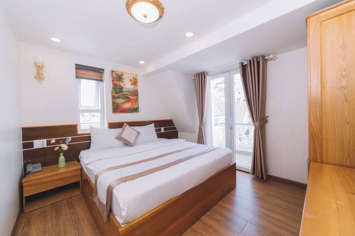 Hotelkamer met een groot bed, houten hoofdeinde, nachtkastje met een vaas, een schilderij aan de muur en uitzicht door het raam.