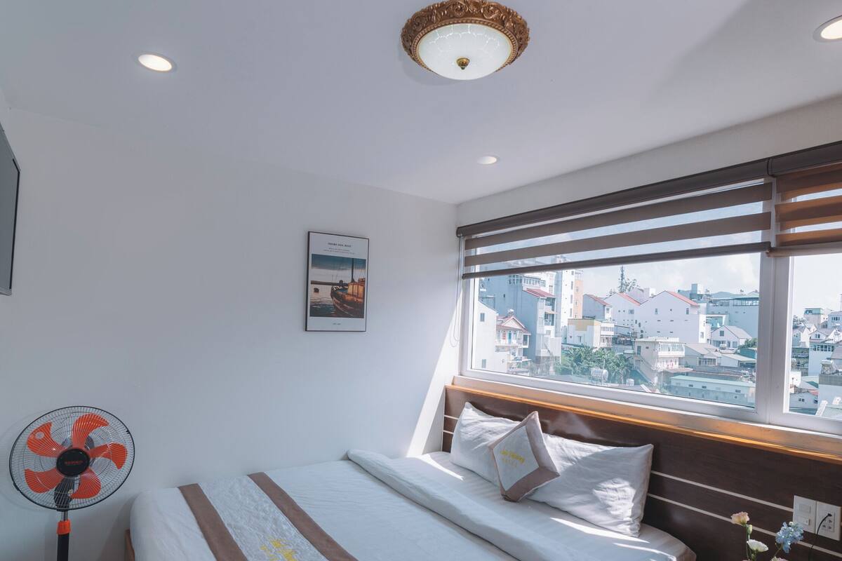 Een slaapkamer met een bed, een ventilator, een raam met uitzicht op gebouwen en een plafondlamp.