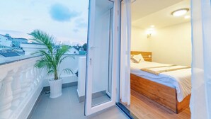 Un balcon agrémenté d’un palmier en pot, une porte-fenêtre coulissante donnant sur une chambre avec un lit en bois et du linge de lit blanc.
