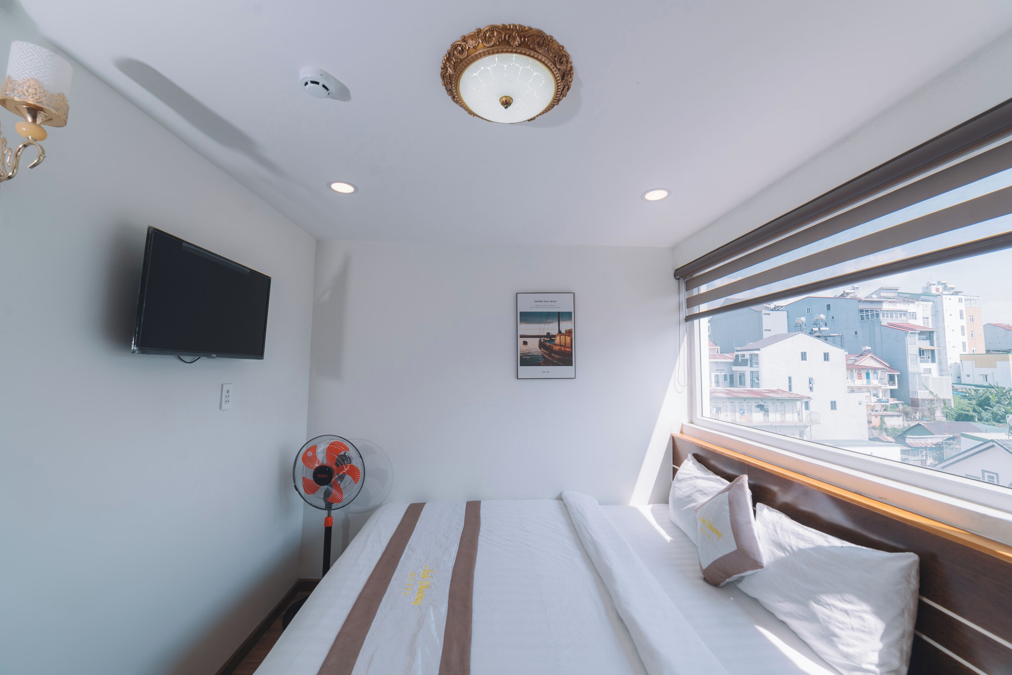 Una habitación de hotel con cama, televisor, ventilador y ventana con vista a la ciudad.