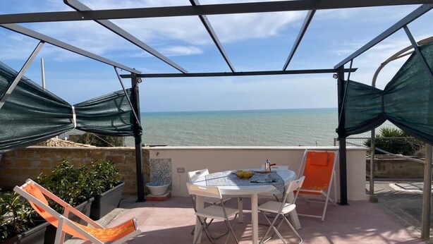 A covered outdoor seating area with a table and chairs, overlooking a body of water. - Cassiopeia House (Porto Palo)
