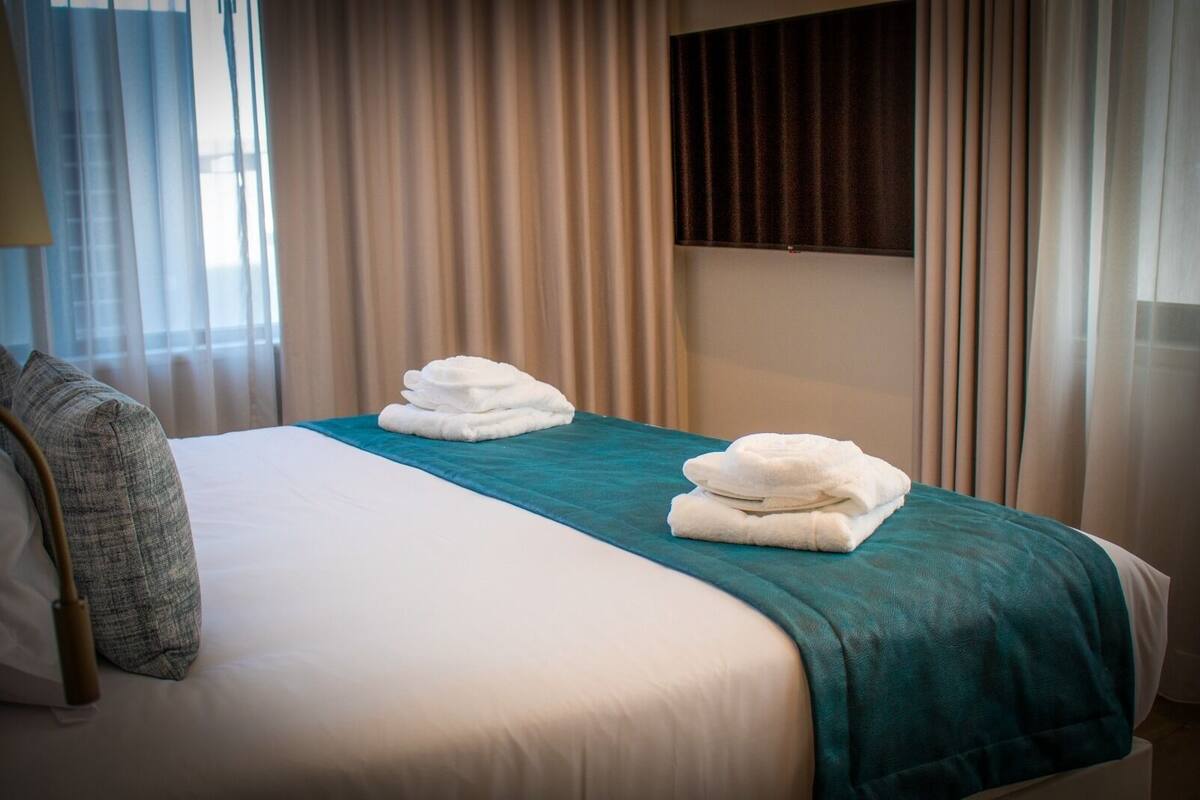 Una habitación de hotel con una cama bien hecha, un plaid verde azulado y dos toallas dobladas sobre la cama.