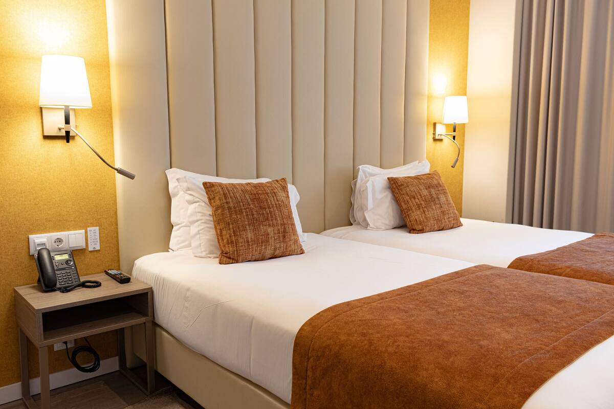 Habitación de hotel con dos camas, una mesita de noche con teléfono y control remoto, y lámparas de pared.