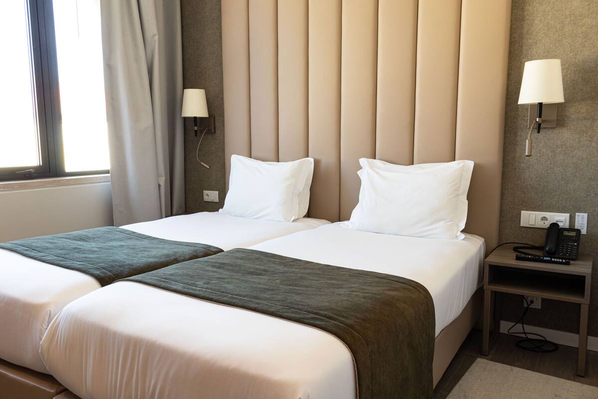 Una habitación de hotel con dos camas, una mesita de noche con teléfono y control remoto, y una ventana con cortinas.