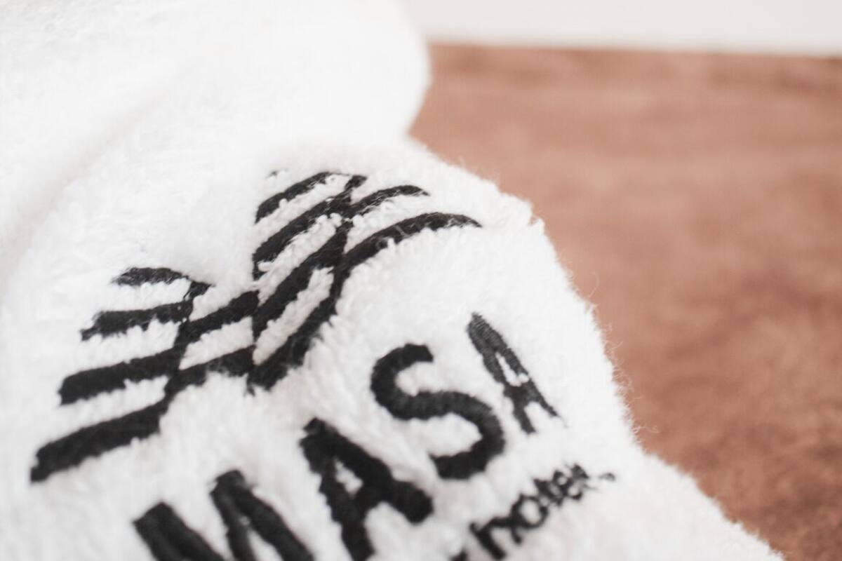 Una toalla blanca con el texto "MASAI" y "grupo o hotel", una cama blanca y una almohada marrón en una habitación de hotel.