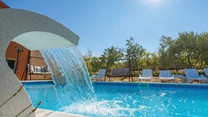 A swimming pool with a water feature, surrounded by lounge chairs and trees.