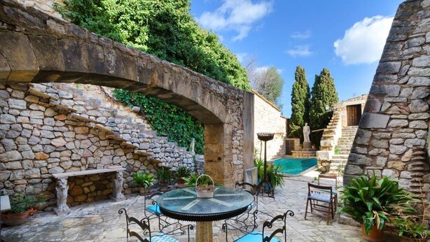 Un patio con paredes de piedra, una mesa redonda y sillas, un banco de piedra y una pequeña piscina.