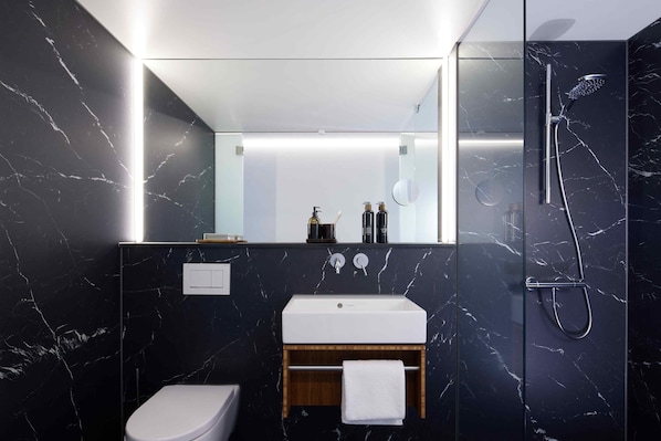 A modern bathroom with a marble wall, a white sink, a wooden vanity, a toilet, and a glass-enclosed shower area.