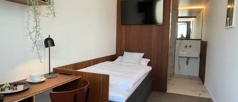 Quarto de hotel compacto com cama, escrivaninha com cadeira, TV fixada na parede e banheiro pequeno com pia e espelho.