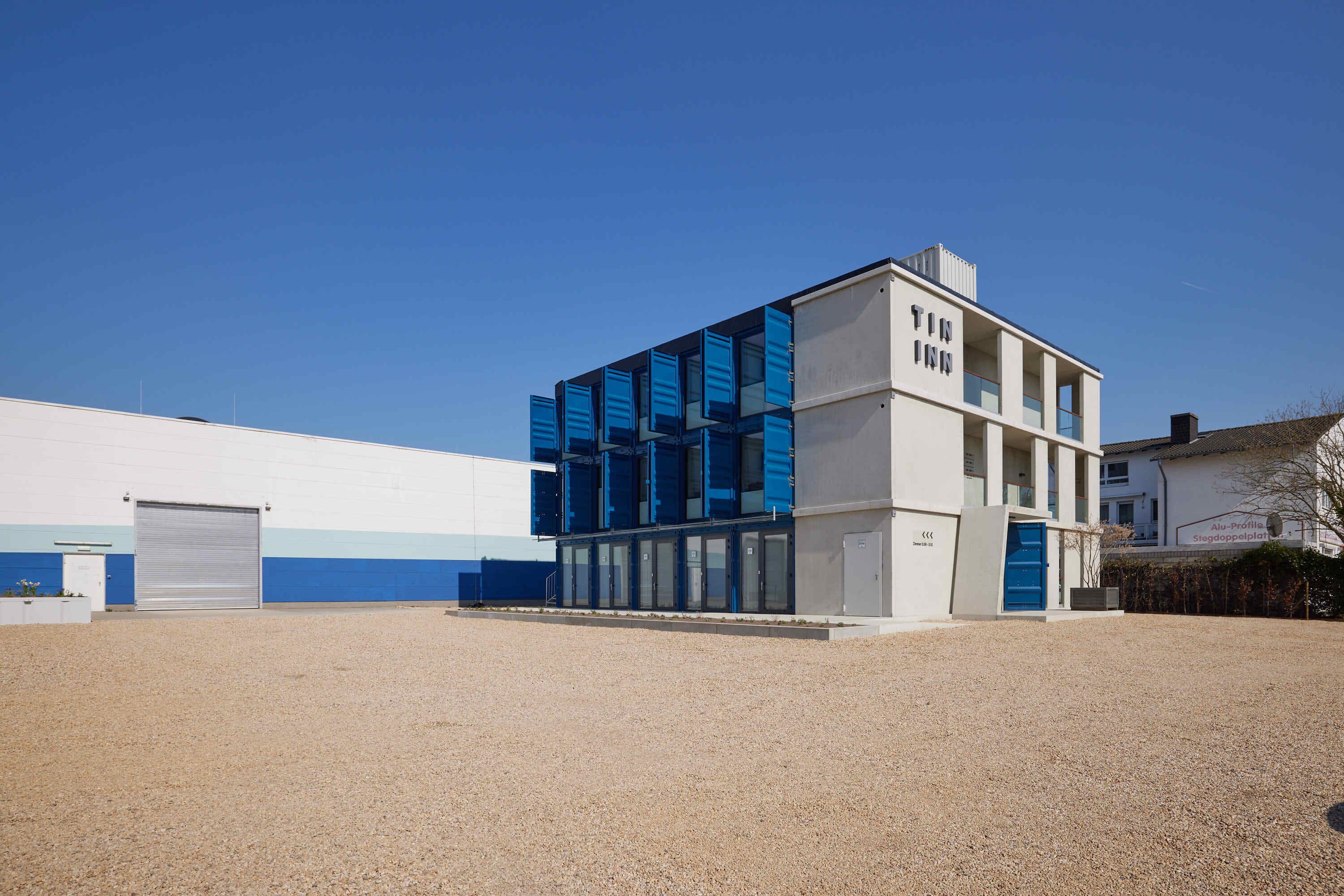Ein modernes Gebäude mit weißer Fassade und blauen Akzenten, mit einer großen Garagentür und einem Schotterweg.