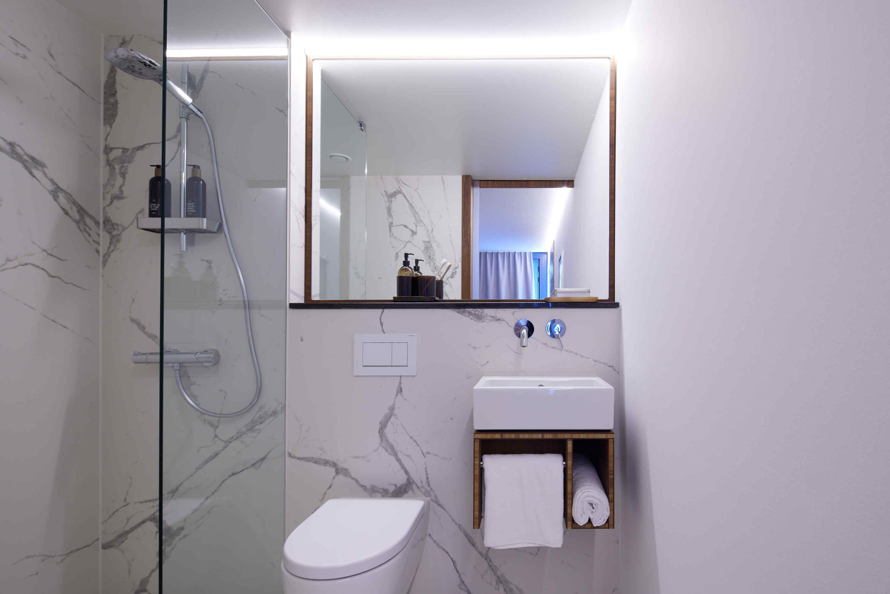 A modern bathroom with a glass-enclosed shower, a white toilet, a marble-tiled wall, a white sink, and a mirror above the sink.
