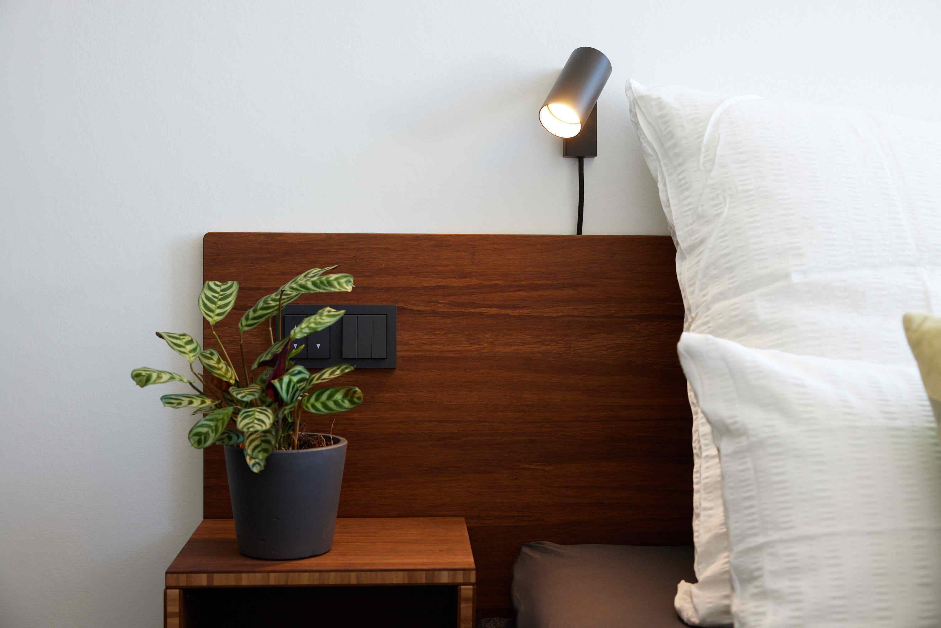 Una mesita de noche de madera con una planta en maceta, una lámpara de pared y un almohadón blanco.