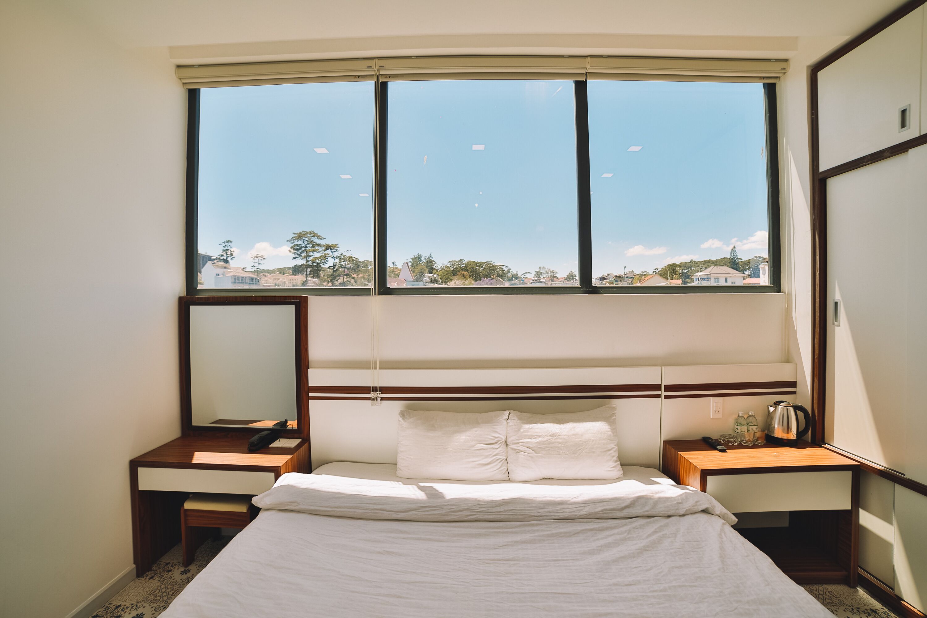 A hotel room with a large window, a bed, a bedside table, a mirror, and a nightstand.