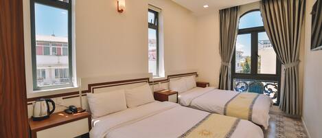 A hotel room with two beds, a window with curtains, a view of buildings outside, and a small balcony with a railing.