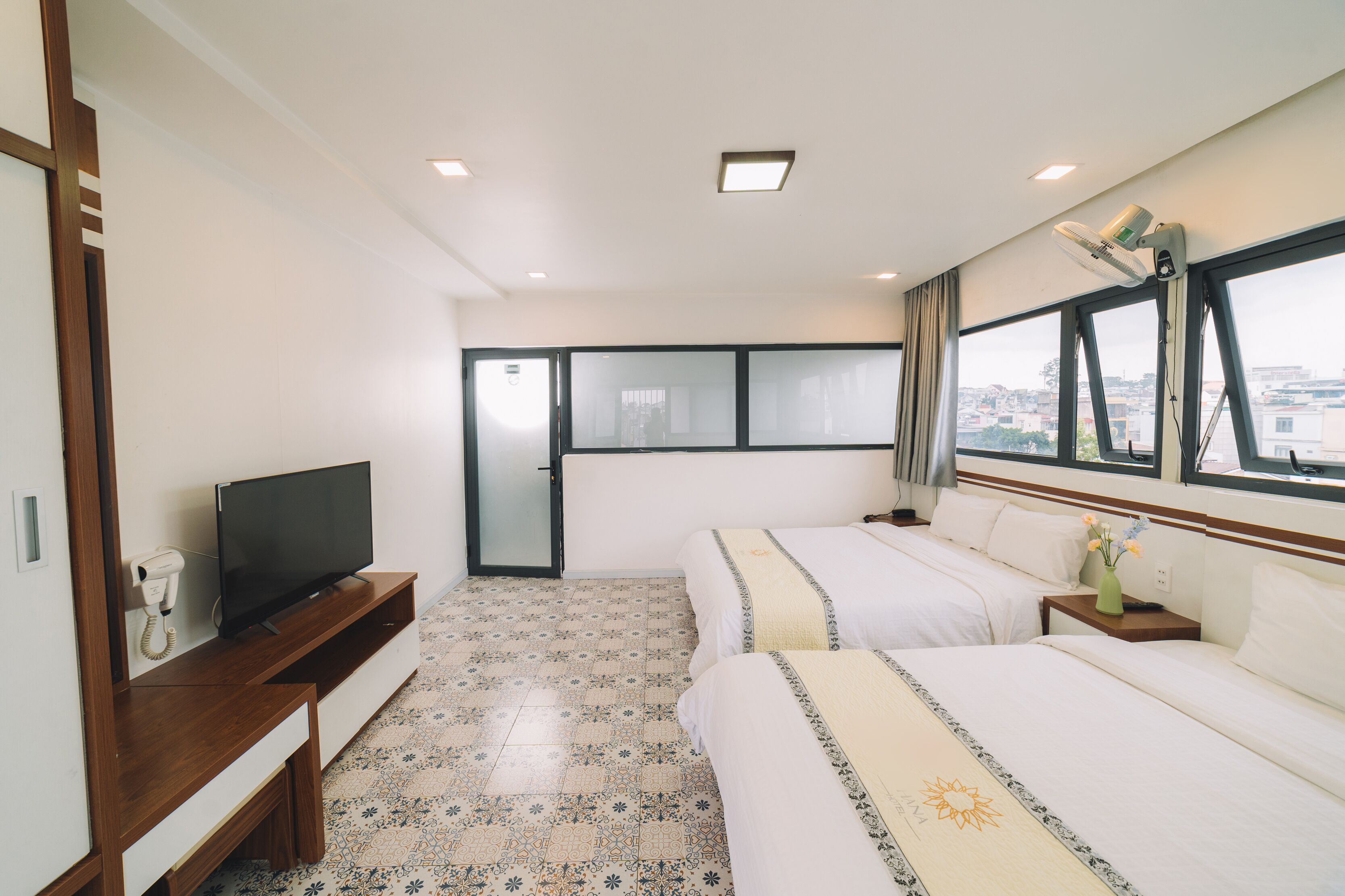 A hotel room with a TV, two beds, a desk, a chair, a window with a city view, and a patterned floor.