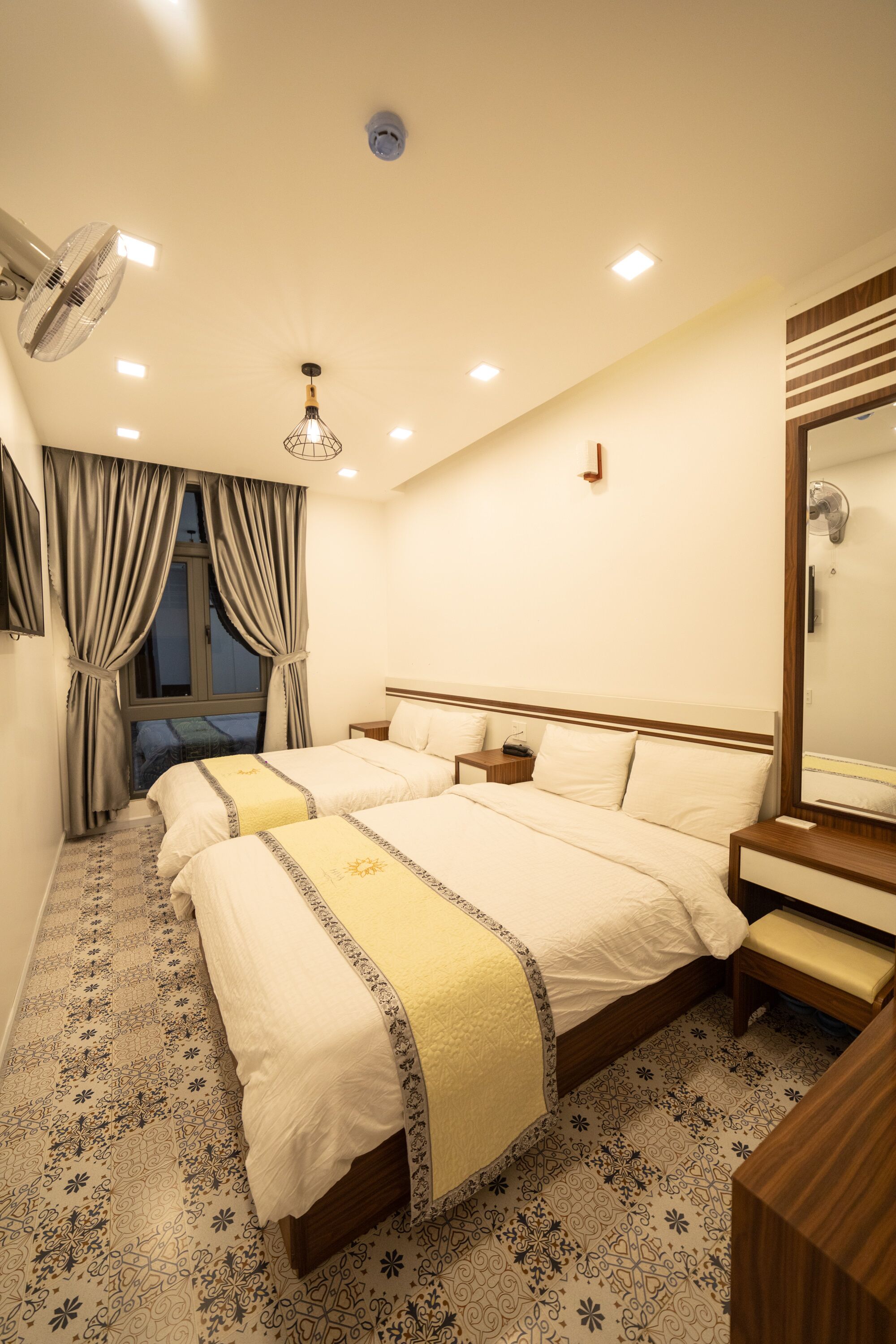 A hotel room with two beds, a large mirror, a bedside table, a nightstand, a ceiling fan, and a window with curtains.
