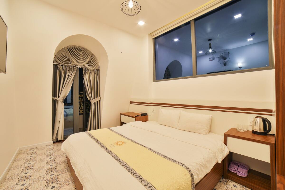A hotel room with a bed, a nightstand, a window with curtains, a ceiling fan, and a view of the outside.