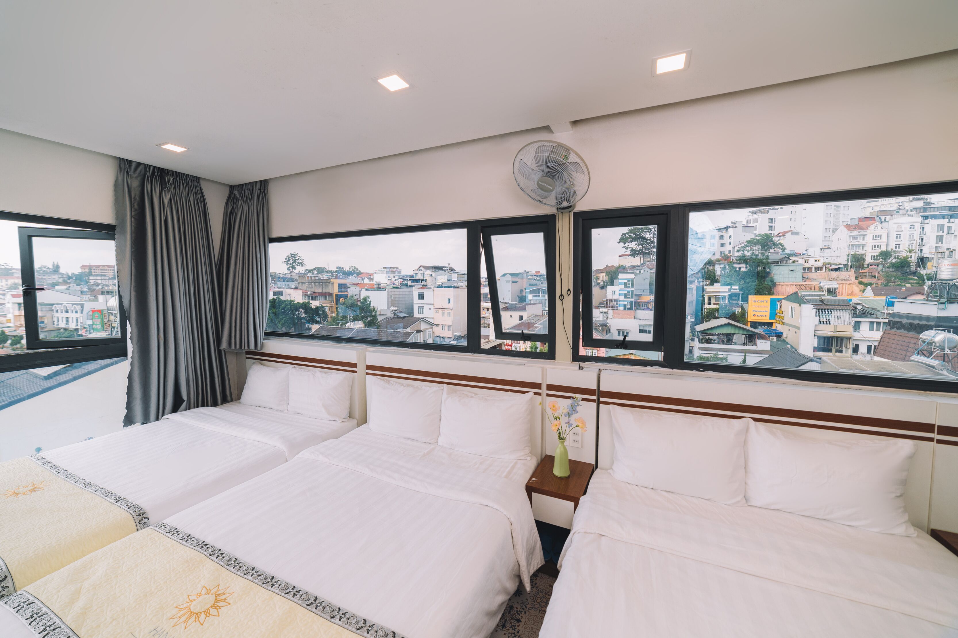 Habitación de hotel con dos camas, un ventanal grande con vista a la ciudad, un ventilador de techo y una mesita de noche con un florero.