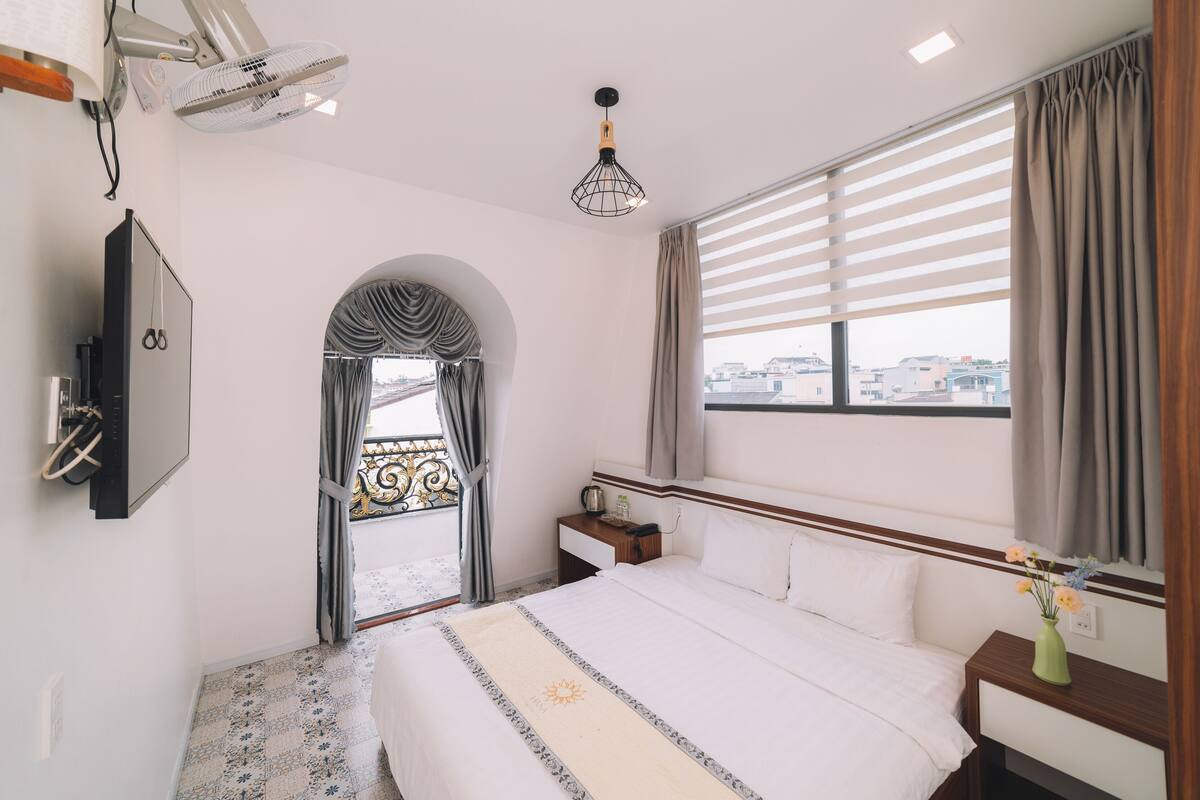 A hotel room with a bed, bedside table, lamp, and a view of the city through the window.