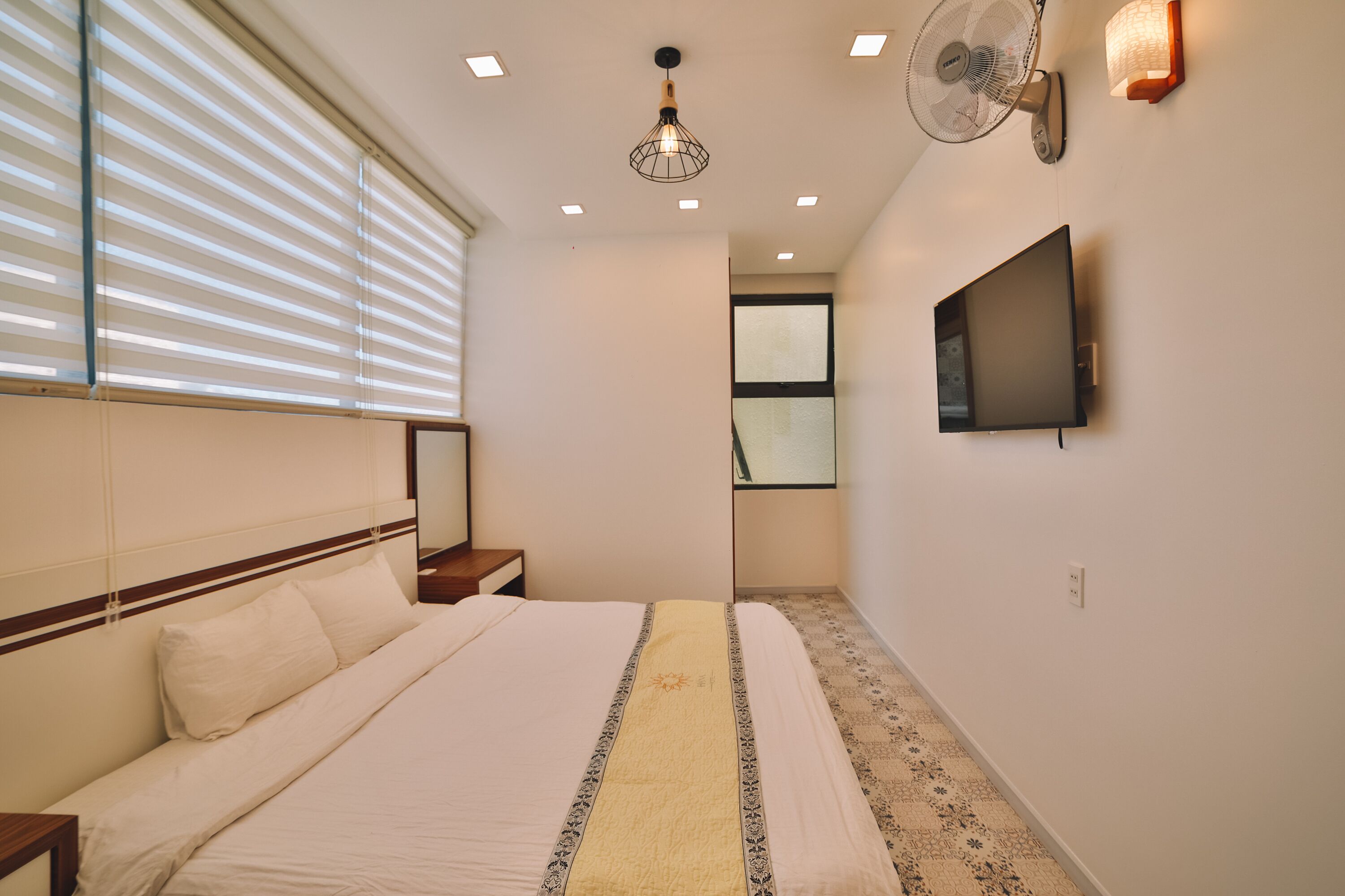 Un dormitorio con cama, mesita de noche, televisor de pared, ventilador de techo y persianas.
