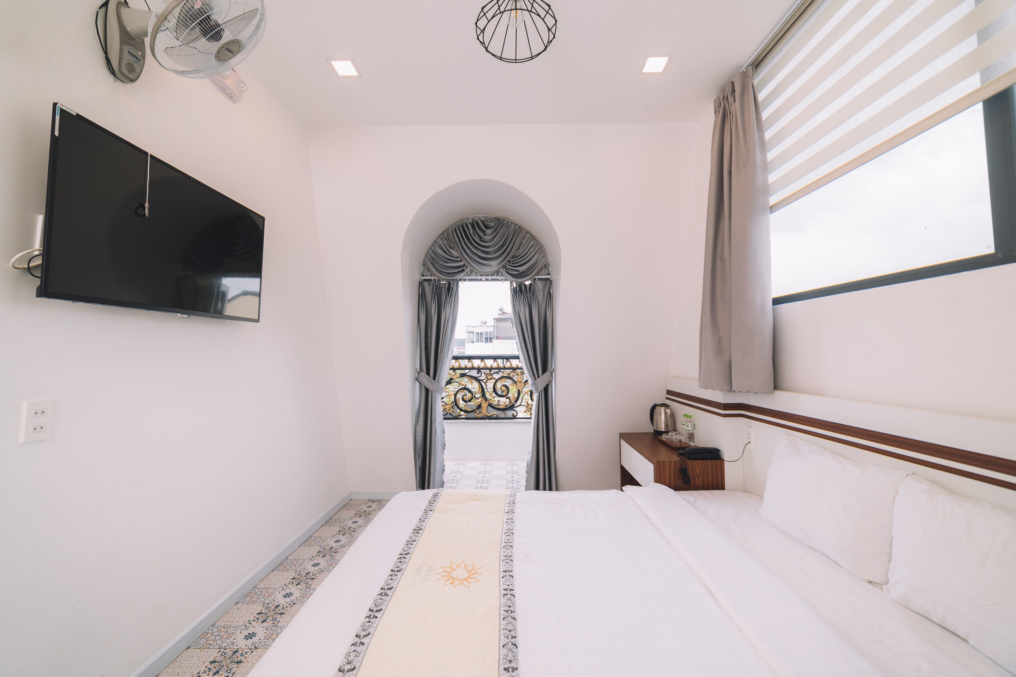 A hotel room with a bed, a TV mounted on the wall, a ceiling fan, and a window with curtains.