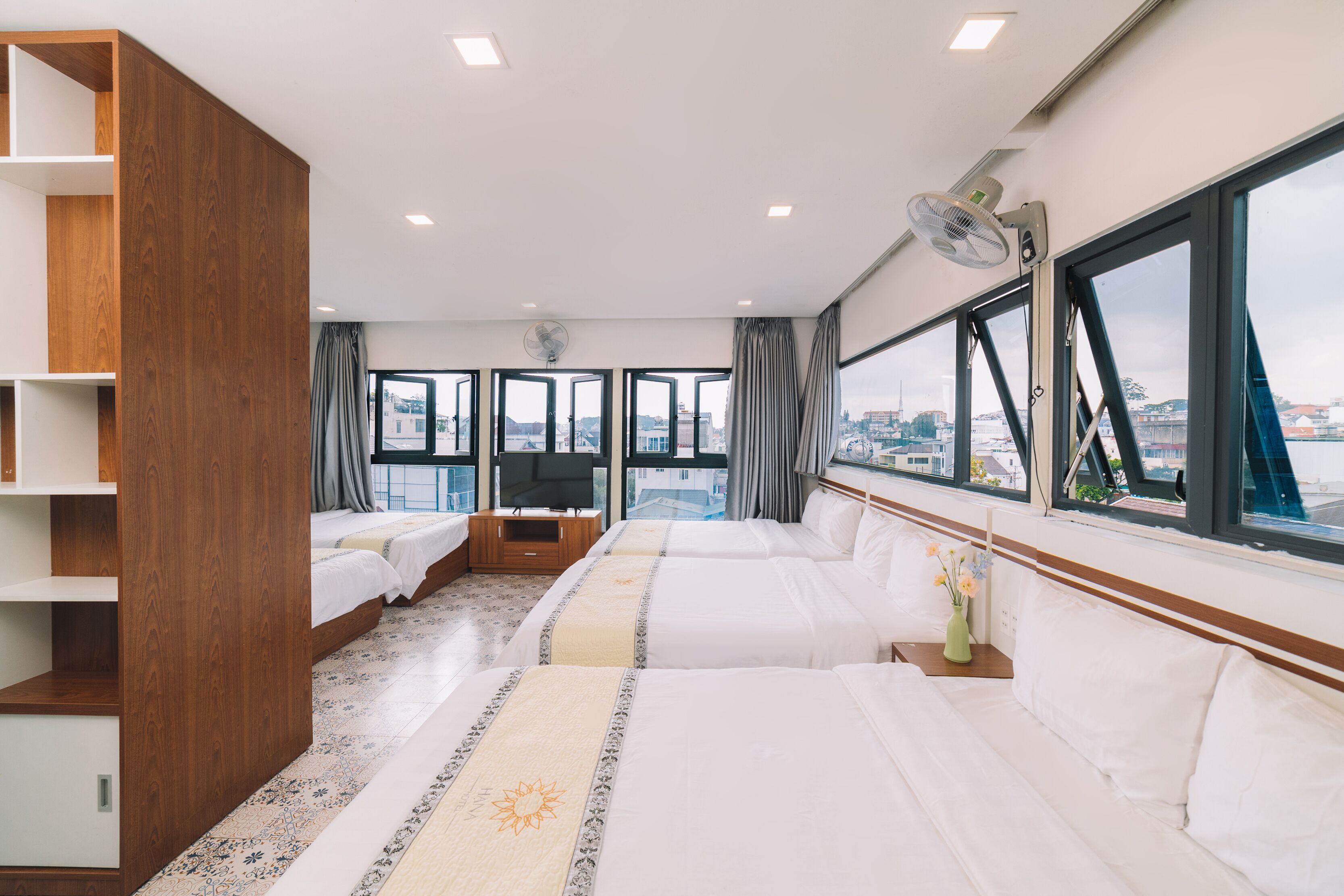 A hotel room with two beds, a large window with a city view, a wooden wardrobe, and a TV on a wooden stand.