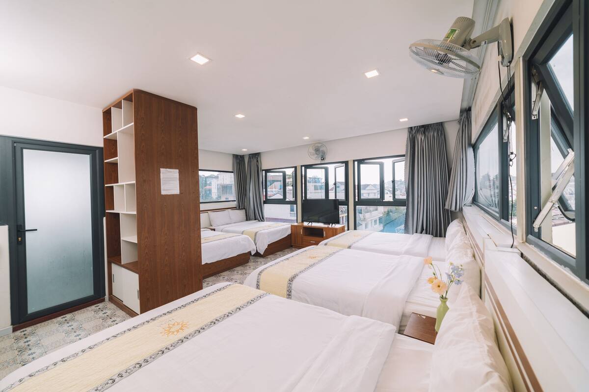 A hotel room with three beds, a wardrobe, a TV, and large windows with curtains.