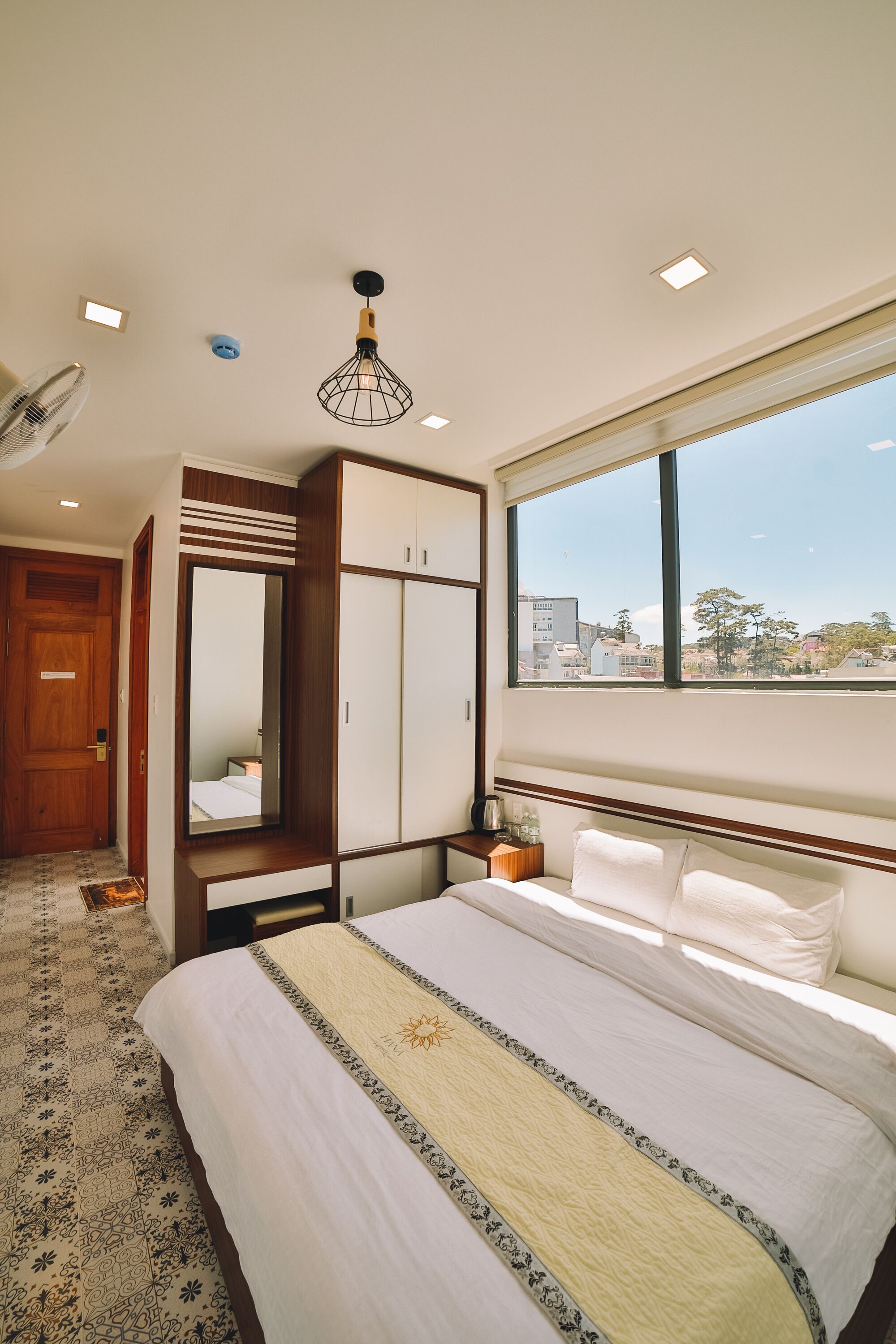 A hotel room with a large bed, a wardrobe, a mirror, a ceiling fan, and a window with a city view.