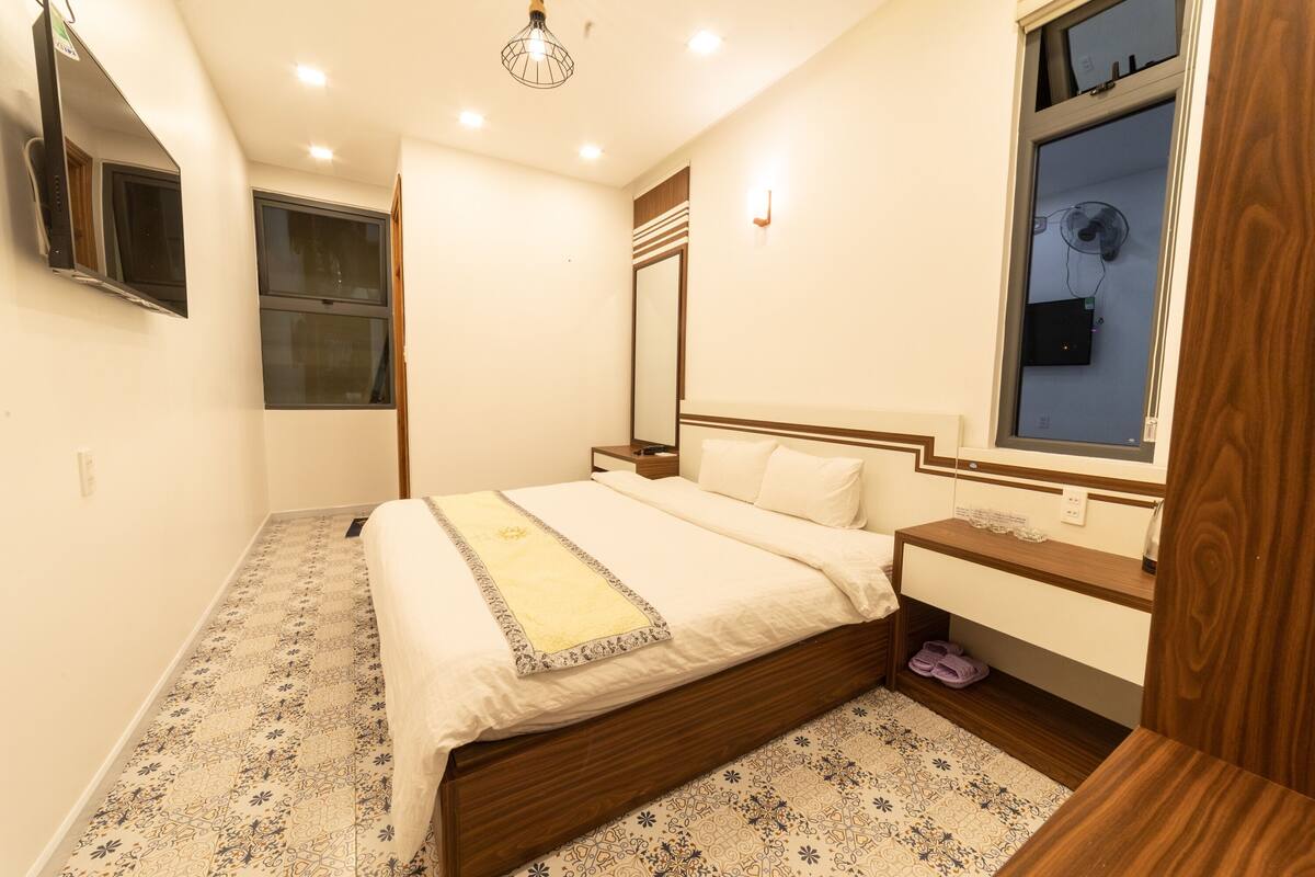 A bedroom with a bed, bedside table, nightstand, and a window with a view.