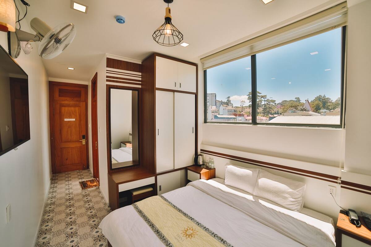 A hotel room with a bed, a wardrobe, a bedside table, a nightstand, a window with a view, and a ceiling fan.