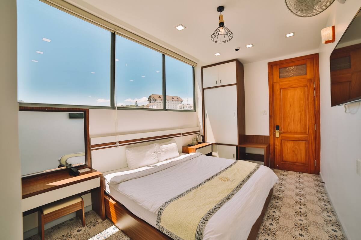 A bedroom with a large window, a bed, a desk, and a wardrobe.