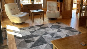 Een gezellig houten blokhutinterieur met twee witte leren fauteuils, een houten tafel en een vloerkleed met dessin.