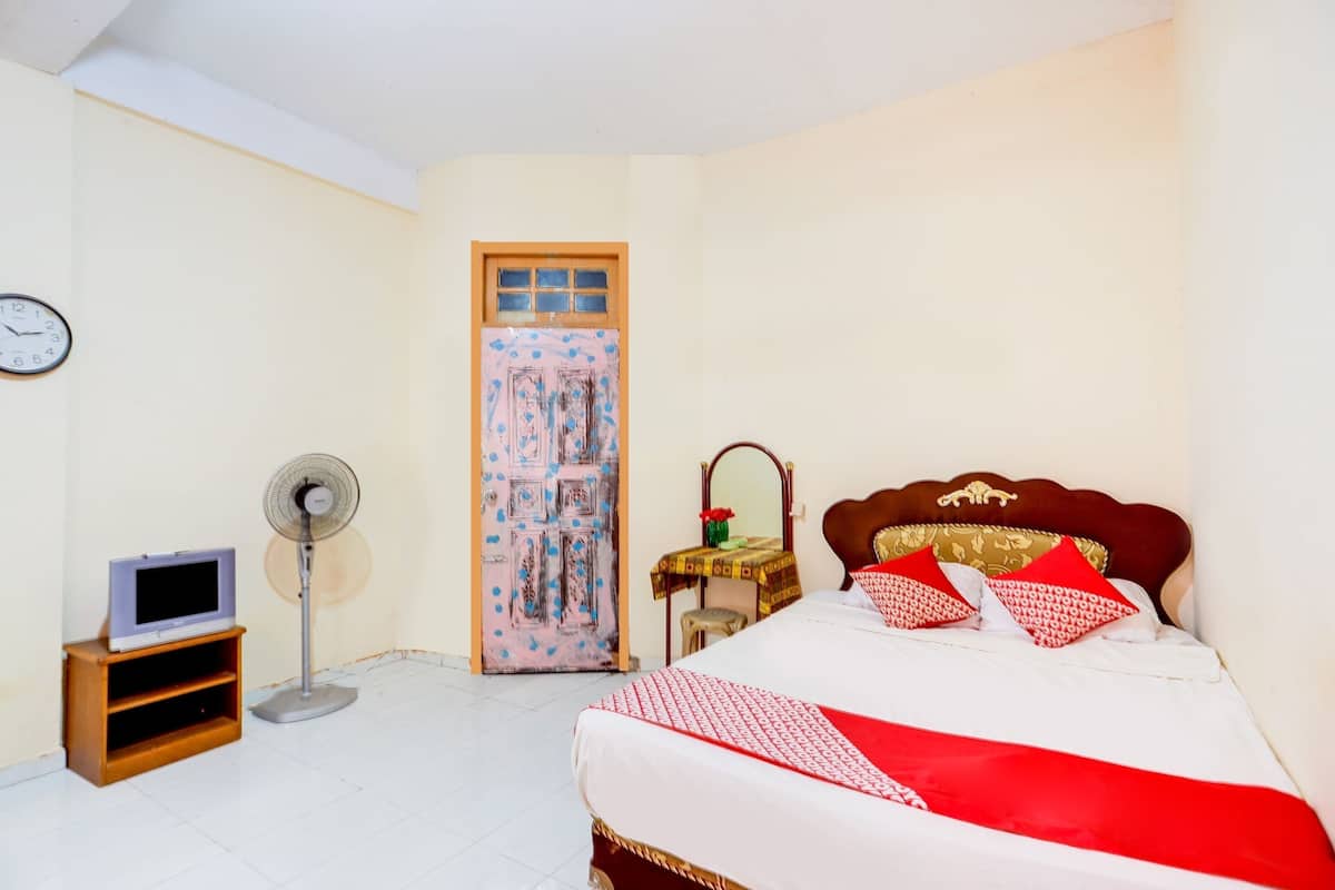 Un dormitorio con cama, televisor sobre un mueble, ventilador, puerta con ventana y un reloj en la pared.