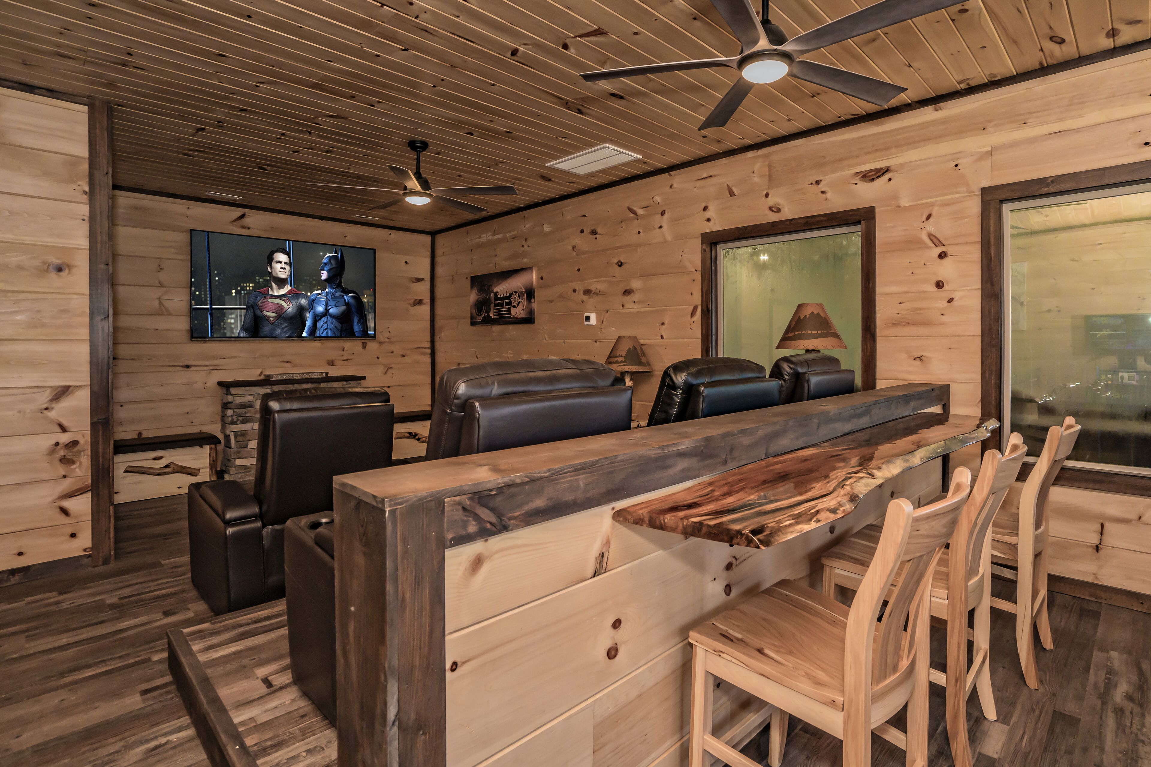 A wooden interior with a ceiling fan, leather seating, a TV displaying superheroes, and a large window.