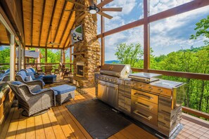 A covered outdoor area with a stone fireplace, a stainless steel grill, wicker furniture, and a view of the forest. - WildernessSplashLodge: Pool, Mtn View, Pets, Theater, Game Room, Hot Tub, WiFi (Sevierville)