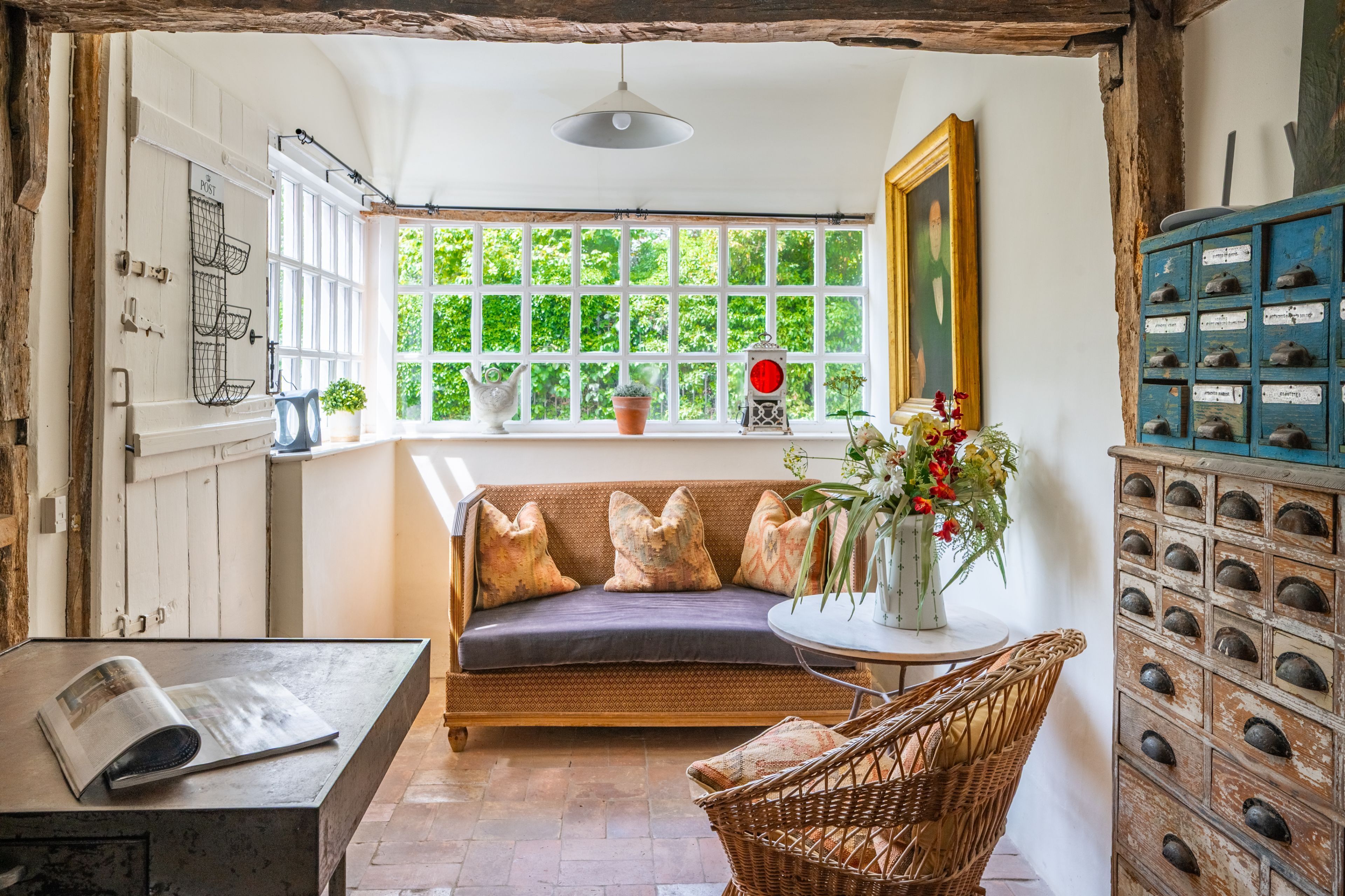 Ein gemütliches Zimmer mit einer Holzbank, einem Korbstuhl, einem kleinen Tisch mit einem Buch, einem Fenster mit Grünpflanzen draußen und einem altmodischen Schrank mit Schubladen.
