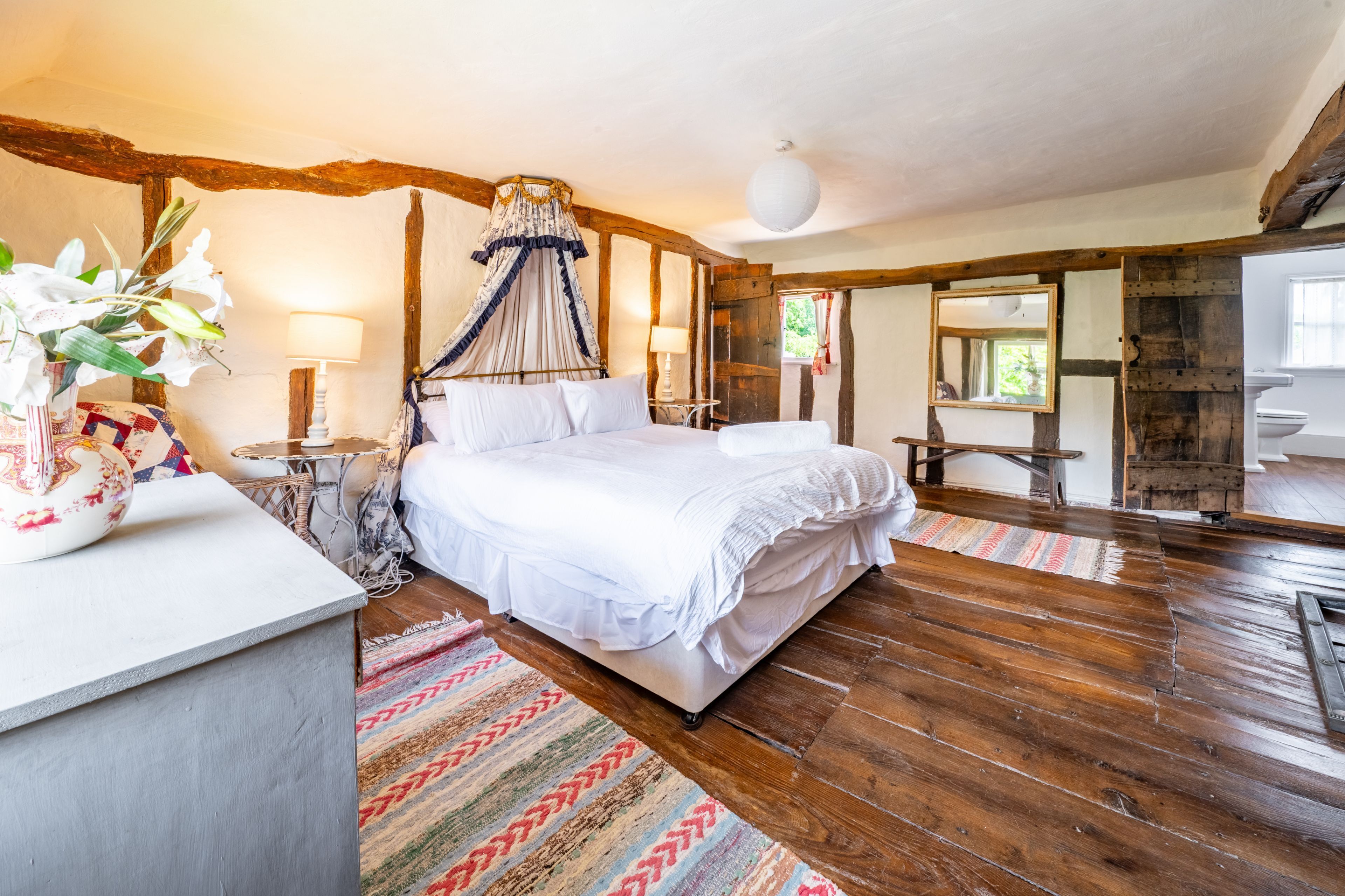 Une chambre à coucher avec un grand lit, une table de chevet avec une lampe, un plancher en bois et une fenêtre avec des rideaux.