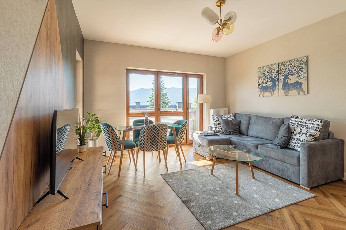 A modern living room with a sofa, a dining table with chairs, a TV, and a large window with a view of mountains.
